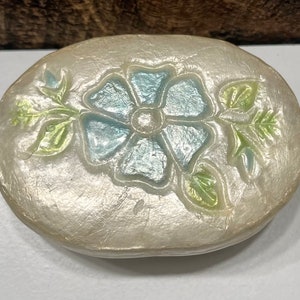 May include: A white oval trinket box with a raised blue flower design and green leaves. The box has a pearlescent finish.