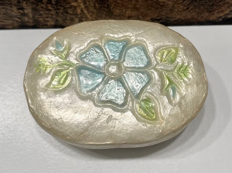 May include: A white oval trinket box with a raised blue flower design and green leaves. The box has a pearlescent finish.