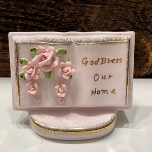 May include: A pink ceramic book with a gold trim and a floral design. The book says "God Bless Our Home" in gold lettering.
