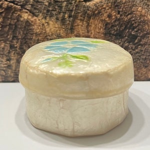 May include: A small, white ceramic trinket box with a blue and green floral design on the lid.