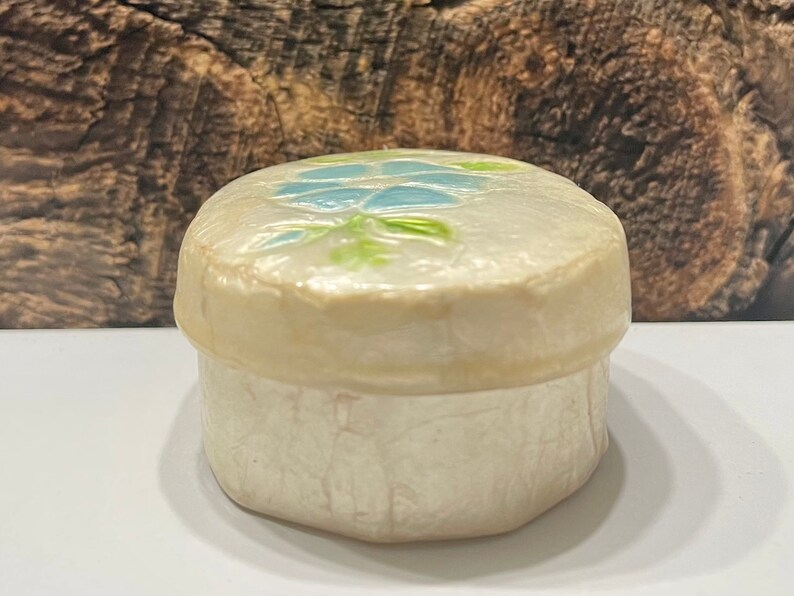 May include: A small, white ceramic trinket box with a blue and green floral design on the lid.