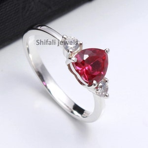 May include: A silver ring with a red heart-shaped gemstone and two smaller white gemstones. The ring is on a white background. Shifali Jewels is written on the band.