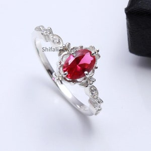 Op de afbeelding: Een zilveren ring met een grote, ovale robijn die is gezet in een delicaat, bloemig ontwerp. De ring is versierd met kleinere, witte edelstenen.