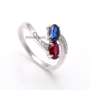 Może przedstawiać: Srebrny pierścionek o nowoczesnym designie z dwoma owalnymi kamieniami szlachetnymi, jednym niebieskim i jednym czerwonym. Obręcz pierścionka ma zakrzywiony kształt, ozdobiona małymi, przezroczystymi kamieniami szlachetnymi. Pierścionek jest umieszczony na białym tle.