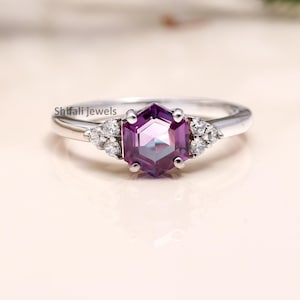 Peut inclure: Une bague en argent ornée d'une pierre précieuse hexagonale violet foncé en son centre. De petits groupes de pierres blanches flanquent la pierre centrale. La bague est posée sur un fond neutre et doux, mettant en valeur le design.