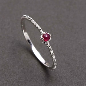 May include: A silver ring featuring a small, round, deep red gemstone. The band has a textured, beaded design on one side. The ring is set against a dark grey background.
