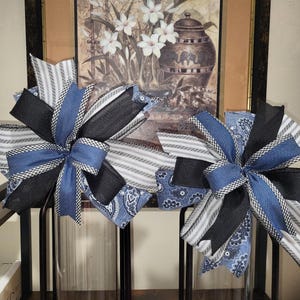 May include: Two decorative bows with a mix of blue, black, and white patterned ribbons. The bows feature denim, striped, and paisley designs, creating a layered and textured appearance. The bows are likely for home decor or gift wrapping.