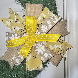 May include: A decorative wreath bow featuring a mix of ribbons in yellow, white, and burlap. The bow has a yellow polka dot ribbon at the center, with daisy and bee patterns on the other ribbons. The bow is attached to a green wreath.