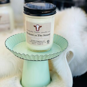 May include: A white candle jar with a black lid and a cow logo. The label reads "uniquely Texan candle co. Dickens on The Strand cinnamon, clove, nutmeg juniper, musk soy wax, fragrance oil hand poured in McGregor, TX approx. 170g 6 oz."