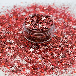 May include: A clear jar filled with red and silver glitter, with a snowflake design. The glitter is scattered around the jar on a white surface.
