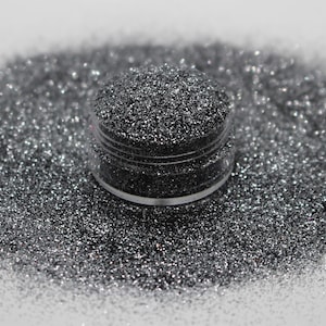 May include: A small clear jar filled with silver glitter. The glitter is spread out around the jar on a white surface.