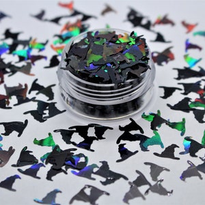 May include: A small jar filled with black iridescent glitter shaped like witch hats. The glitter is scattered around the jar on a white surface.