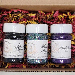 Puede incluir: Tres pequeños frascos de purpurina, cada uno con un color y una etiqueta diferentes. Los frascos están etiquetados como "Be Witched", "Hocus Pocus" y "Purple Rain".