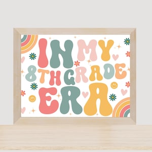 May include: A framed print with a white background and colorful text that reads "In My 8th Grade Era". The text is in a retro font style and is surrounded by flowers, hearts, and rainbows.