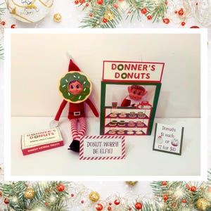 May include: A Christmas elf wearing a green and white donut costume sits in front of a red and green cardboard donut shop stand. The shop sign reads "Donner's Donuts". A sign below the shop reads "Donut Worry! Be Elfie!" and another sign reads "Donuts £1 each or 12 for £10".