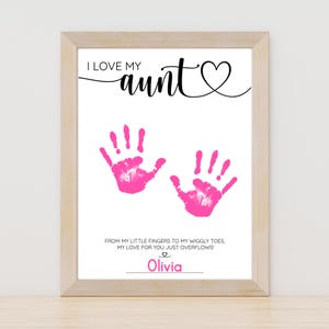 May include: Framed print with the text "I LOVE my aunt" in black script, featuring two pink handprints. Below, the text reads "FROM MY LITTLE FINGERS TO MY WIGGLY TOES, MY LOVE FOR YOU JUST OVERFLOWS!" with the name "Olivia" below.