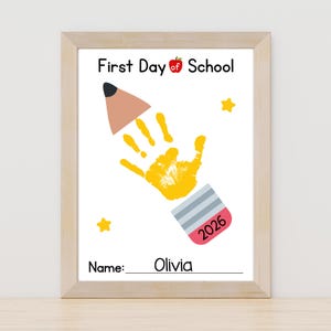 Puede incluir: Una impresión enmarcada para el primer día de clases, con una huella de mano amarilla que forma un lápiz. La impresión incluye el texto "First Day of School", una manzana, estrellas y el nombre "Olivia". También es visible el año "2026".