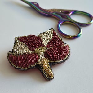 Peut inclure: Gros plan d'une broche faite à la main en forme de feuille brodée avec du fil rouge et doré. La broche est à côté d'une paire de ciseaux à broder irisés. La broche a une bordure noire.