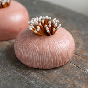 May include: Two pink ceramic match holders. Each holder is topped with a cluster of matches with white tips. The holders sit on a weathered wooden surface. The holders have a textured surface.