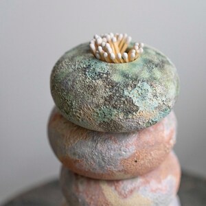 May include: A stack of four ceramic rings in muted colours, topped with a cluster of wooden matches. The top ring is a mottled green, while the others transition from pink to blue. A unique home decor item.