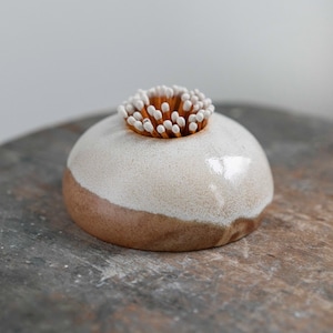 May include: A ceramic match striker with a rounded form, in a two-tone design. The top half is creamy white, the bottom a warm brown. A cluster of wooden matches is inserted in the top. The item is designed for striking matches.