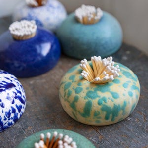 May include: Several ceramic match holders in shades of blue, green, and yellow. Each round holder has a central opening filled with matches. The holders feature speckled or solid glaze finishes, displayed on a wooden surface.
