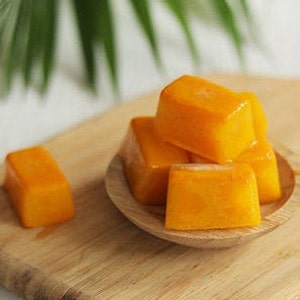 May include: A small wooden bowl filled with bright orange mango cubes. The bowl is sitting on a light brown wooden surface.