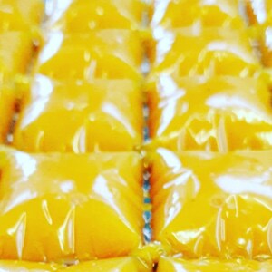 May include: A close-up of a tray of individually packaged yellow squares of a sweet treat. The squares are arranged in rows and are wrapped in clear plastic.