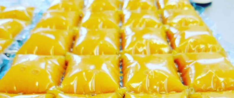 May include: A close-up of a tray of individually packaged yellow squares of a sweet treat. The squares are arranged in rows and are wrapped in clear plastic.