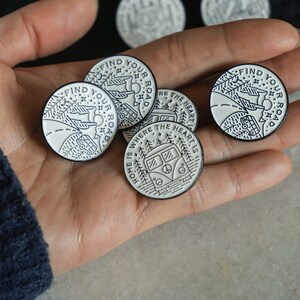 May include: Five enamel pins with white backgrounds and black outlines. The pins feature illustrations of a van, trees, and mountains. The text on the pins reads "Find Your Road" and "Home is Where the Heart Is".