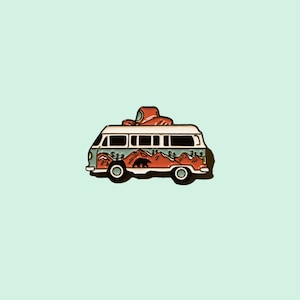 May include: A colorful enamel pin featuring a vintage camper van with a mountain and bear design. The van is painted in shades of green, orange, and white, with a red object on top. The pin is set against a light blue background.