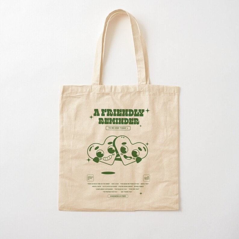 May include: A beige tote bag with a green design that says "A Friendly Reminder to be kind today." Two green cartoon hearts with faces are holding hands. The text "Kindness is free" is at the bottom of the bag.