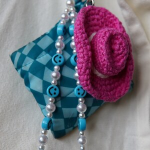 K&ouml;nnte beinhalten: Ein Schl&uuml;sselanh&auml;nger mit einem geh&auml;kelten rosa Cowboyhut, einem t&uuml;rkis-wei&szlig; karierten Quadrat und einer Perlenkette. Die Kette hat wei&szlig;e Perlen und blaue Perlen mit Smiley-Gesichtern.