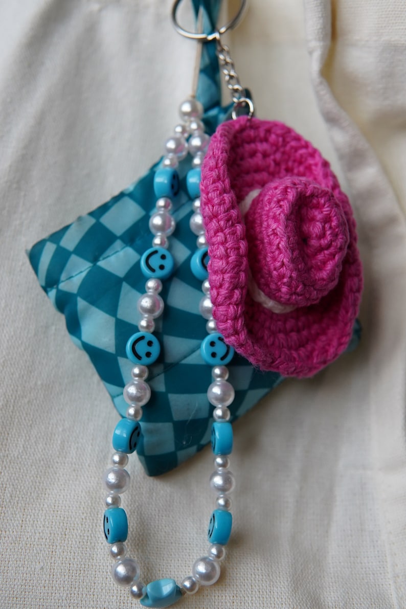 K&ouml;nnte beinhalten: Ein Schl&uuml;sselanh&auml;nger mit einem geh&auml;kelten rosa Cowboyhut, einem t&uuml;rkis-wei&szlig; karierten Quadrat und einer Perlenkette. Die Kette hat wei&szlig;e Perlen und blaue Perlen mit Smiley-Gesichtern.