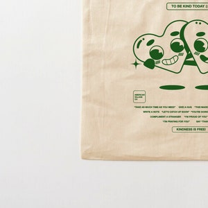 May include: A beige tote bag with a green design of two cartoon hearts holding hands. The text "TO BE KIND TODAY :)" is at the top of the bag. The text "KINDNESS IS FREE!" is at the bottom of the bag. The text "AMERICAN VILLAGE CO" is in the center of the bag.