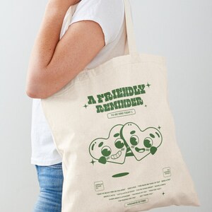 May include: A white tote bag with green text that reads "A Friendly Reminder To Be Kind Today". The bag features a cartoon illustration of two green hearts with faces.