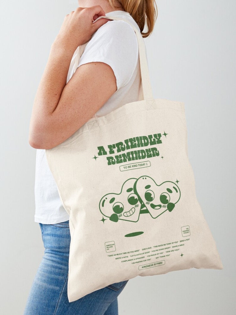 May include: A white tote bag with green text that reads "A Friendly Reminder To Be Kind Today". The bag features a cartoon illustration of two green hearts with faces.