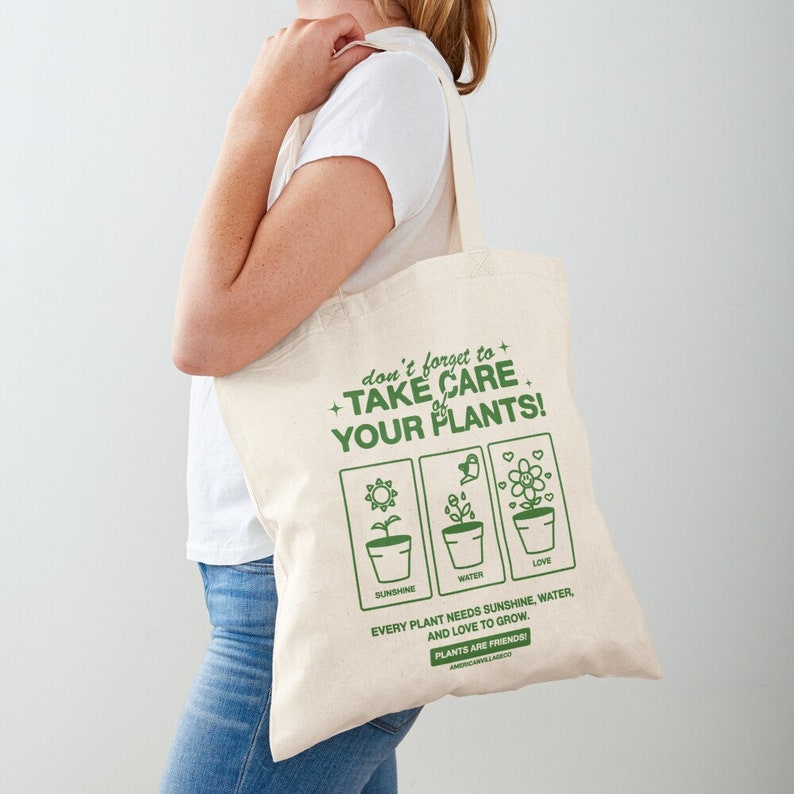 May include: A white tote bag with green text that reads "don't forget to + TAKE CARE of YOUR PLANTS!"  The bag has three green illustrations of potted plants with the words "sunshine", "water", and "love" below each. The text below the illustrations reads "Every plant needs sunshine, water, and love to grow. PLANTS ARE FRIENDS!"
