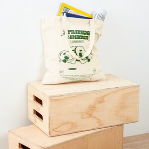 May include: A beige canvas tote bag with green text that reads "Friendly Reminder To Be Kind Today." The bag features a cartoon illustration of two green hearts with eyes and mouths. The bag is sitting on top of two wooden boxes.