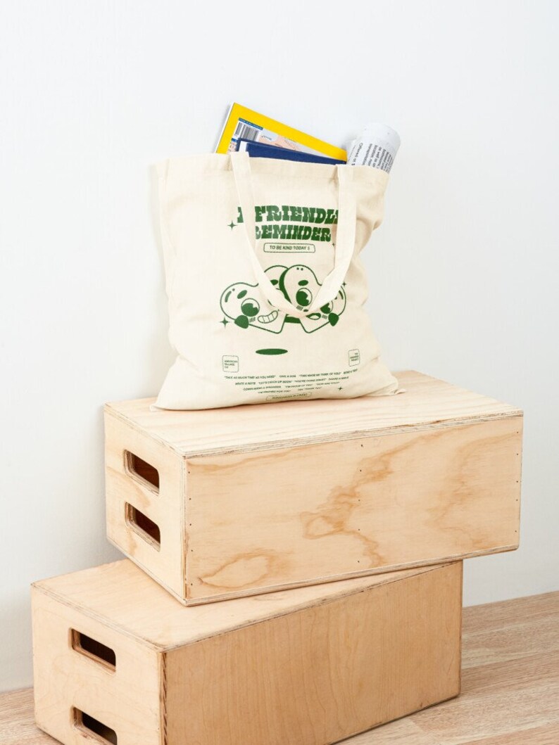 May include: A beige canvas tote bag with green text that reads "Friendly Reminder To Be Kind Today." The bag features a cartoon illustration of two green hearts with eyes and mouths. The bag is sitting on top of two wooden boxes.