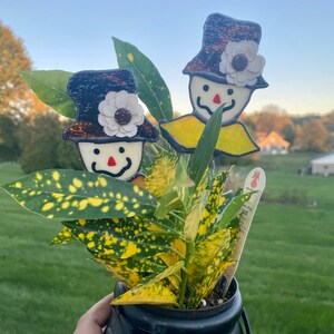 May include: Two glass, stained glass, whimsical, smiling, snowman figures with black hats and white flowers. The figures are in a black pot with a green and yellow variegated plant.