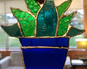 May include: A stained glass succulent plant in a blue pot. The plant has green and blue leaves and is framed in silver.