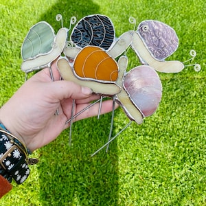 May include: Five stained glass snails in various colors, including green, black, orange, pink, and purple, with silver wire accents. The snails are arranged in a cluster and are attached to metal wires.
