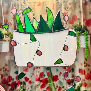 May include: A stained glass hanging featuring a white planter with green leafy plants and pink glass beads hanging from the top.