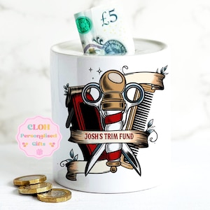 May include: White ceramic money bank with a barber shop design. The bank features a red, white, and gold barber pole, scissors, and a comb. A five-pound note is visible in the slot. The text "JOSH'S TRIM FUND" is on a banner.