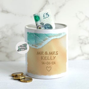 May include: White ceramic piggy bank with a beach scene design. The piggy bank has a slot for coins and says "MR & MRS KELLY 14-06-24" with a heart.