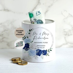 May include: White ceramic piggy bank with a blue and white floral design. The piggy bank has a slot for coins and says "Mr & Mrs Richardson Wedding Fund Engaged 02.02.23" on the front.