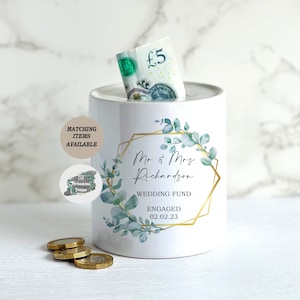 May include: White ceramic wedding fund money box with a gold geometric frame and eucalyptus leaf design. The text reads "Mr & Mrs Richardson WEDDING FUND ENGAGED 02.02.23". A five-pound note is visible in the slot, with coins nearby.