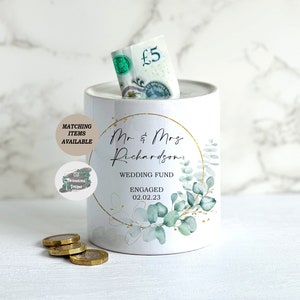 May include: A white cylindrical wedding fund money box with a gold circular design and eucalyptus leaves. The text reads "Mr & Mrs Richardson, Wedding Fund, Engaged 02.02.23." A five-pound note is visible in the slot, with coins in front.