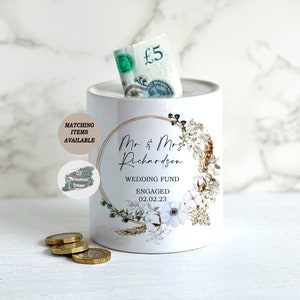 May include: White ceramic piggy bank with a floral design and gold accents. The text "Mr & Mrs Richardson Wedding Fund Engaged 02.02.23" is printed on the front.